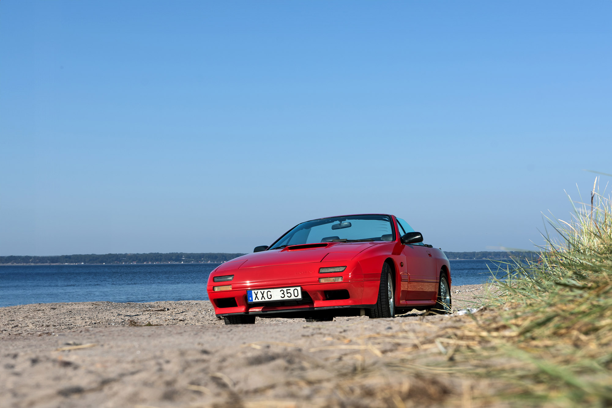 Röd Mazda RX7 parkerad på en sandstrand med blå himmel och hav i bakgrunden, fotograferad i Helsingborg.